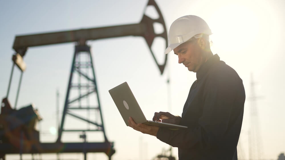 Engineer with a hard hat using a laptop near an oil pump jack.