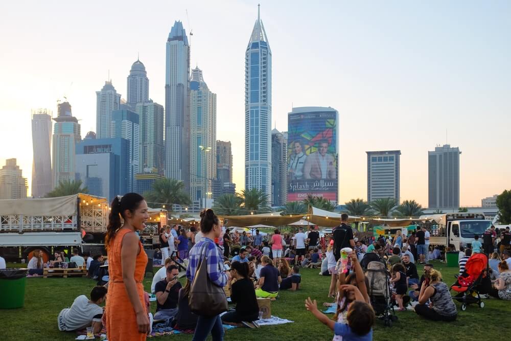 Outdoor market with people relaxing on the grass and Dubai skyline in the background.