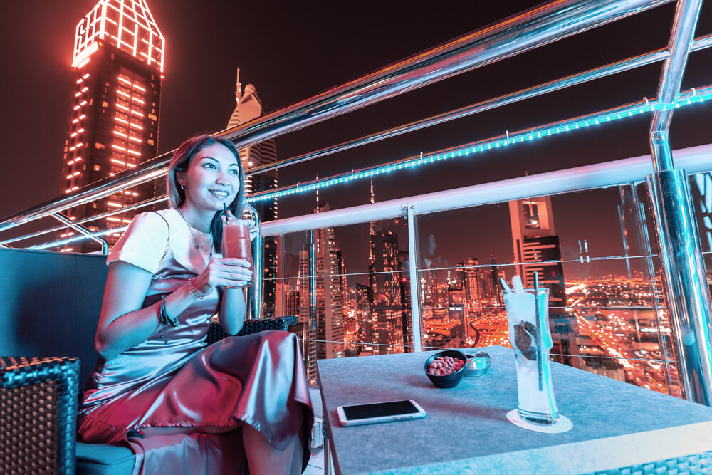 Woman enjoying a drink on a rooftop with the Dubai skyline at night.